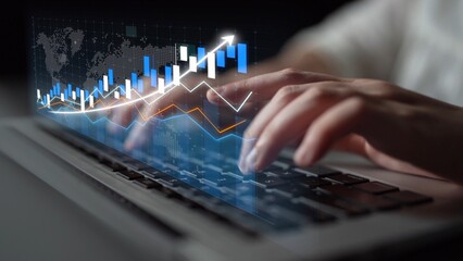 Focused hands typing on a laptop with an overlay of a financial chart, representing the intersection of technology and economic analysis in a modern workspace. Copula