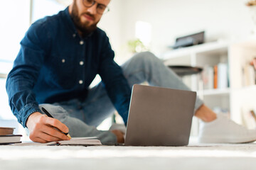 A young man sits comfortably on the floor, focusing on his studies. He uses a laptop placed in front of him and writes notes in a notebook. Sunlight fills the room, creating a warm atmosphere.