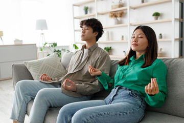 A young Asian couple sits on a couch with closed eyes, meditating together. They practice yoga and relax in a serene living room filled with natural light and greenery for stress relief and wellness.