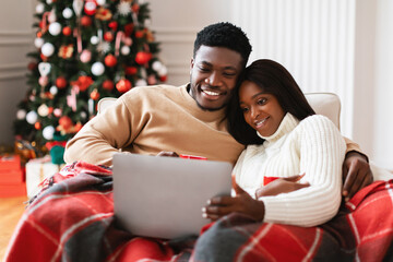 African American couple celebrates Christmas Eve by watching movies on a laptop. They are cozy on...