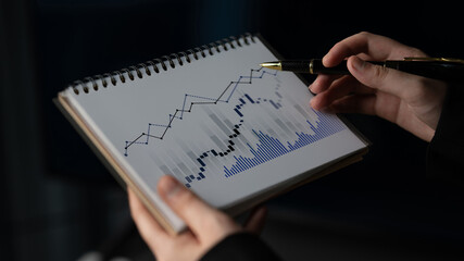A focused investor examines stock market trends using a notebook filled with financial graphs and...