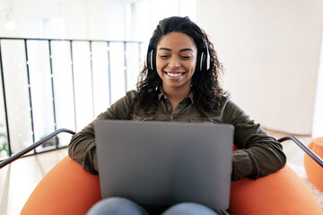 A young woman sits comfortably in an orange chair, using a laptop with headphones on. She smiles...