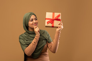 Special occasion concept. Thoughtful arab lady holding wrapped gift, looking at box and thinking...
