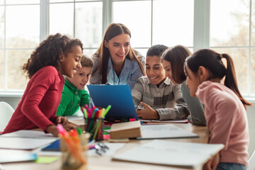 Joyful school children engage with their teacher in a dynamic learning session at their desks. They explore study materials together in a bright, inviting classroom setting.