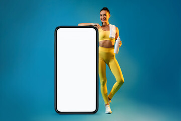 Happy fitness woman in yellow activewear stands by a large smartphone with a blank screen. She...