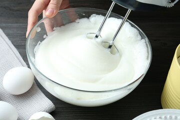 Woman whisking egg whites with hand mixer at black wooden table, closeup
