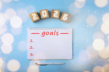New 2026 Year Goals. Inscription in notebook and cubes with numbers on light blue background, top view. Bokeh lights