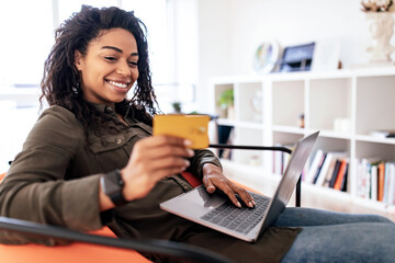 A woman is sitting comfortably in an orange chair, smiling as she holds a credit card in one hand and types on a laptop with the other. The setting is a bright, modern living room.