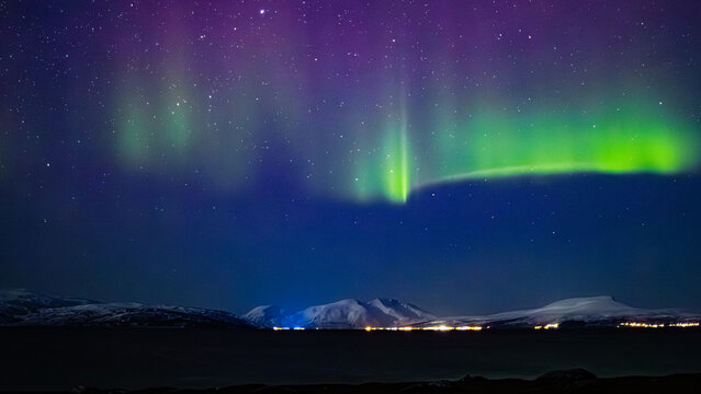 Northern lights or aurora borealis of green and purple color in starry night sky above snow covered mountains and dark water of fjord of tromso norway. City lights shimmering in distant horizon.