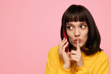 A thoughtful portrait of a young woman in a yellow sweater, listening on a phone while signaling for quiet, set against a soft pink studio background.