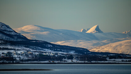 Tromsø, norway winter landscape with small houses on slopes of snow covered mountains near shoreline of fjord. Golden arctic sun light on distant snow capped mountains peaks.
