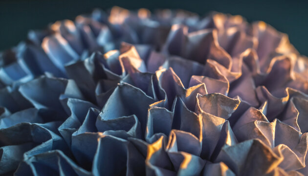Close-up of intricately folded blue and purple paper or fabric with warm light highlighting the textured surface.