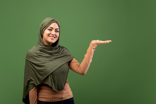 A cheerful arab lady wearing a hijab stands confidently on a dark green background, pointing to the side with her open palm as if presenting something to the audience. She looks friendly and inviting.