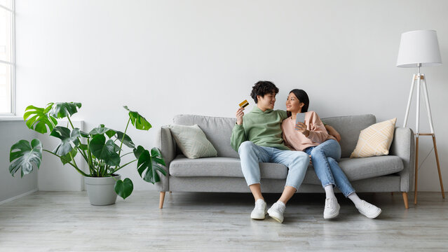 A cheerful young Asian couple sits on their sofa in a bright living room, engaging in online shopping. The man holds a credit card while the woman smiles at him, surrounded by plants and a lamp. - Powered by Adobe