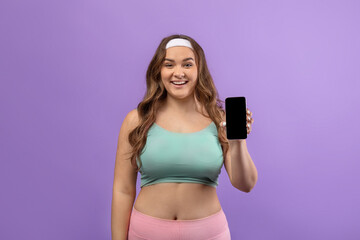 A smiling young European plus size female in sportswear showcases her phone with an empty screen, recommending a new fitness blog.
