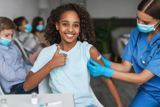 Inside a clinic, a cheerful Black preteen girl smiles and shows a thumbs-up after receiving her coronavirus vaccination.