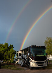 A picturesque RV park is illuminated by a vibrant double rainbow arching across the sky. Trees and a few other campers are visible in the background, creating a serene outdoor atmosphere.