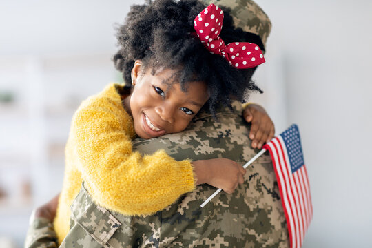 Fototapeta A cheerful young girl with curly hair hugs her father, a soldier, wearing a camouflage uniform. She holds a small American flag and smiles brightly, celebrating their joyful reunion.