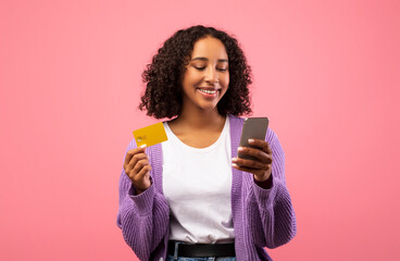 A charming African American woman smiles while shopping online. She holds her smartphone in one...
