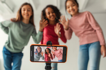 Three multicultural girls are indoors waving to a mobile phone while making a video for their blog....