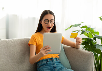 A young woman with glasses shows excitement as she uses a tablet and credit card for online...