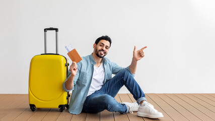A man is seated on the floor next to a bright yellow suitcase, holding his travel documents. He has a big smile and points joyfully, indicating his eagerness to travel.