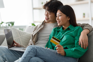 Happy young married Asian couple sits on a couch, holding a credit card and using a laptop. They are shopping online, enjoying their time together in a comfortable home setting.