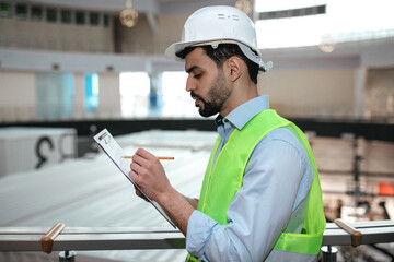 A construction worker wearing a hard hat and safety vest stands on a balcony, reviewing plans and taking notes. The busy construction site is visible in the background.