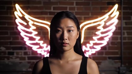 Asian woman poses with glowing neon angel wings on brick wall background for artistic portrait