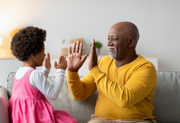 A cheerful little girl and her grandpa share a joyful moment at home, playing a clapping game in their living room. They enjoy their time together, filled with laughter and happiness.