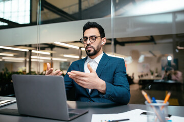 A man dressed in a tailored suit is actively participating in a video call from his stylish office. He appears focused and is using hand gestures to communicate effectively.