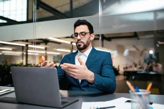 A man dressed in a tailored suit is actively participating in a video call from his stylish office. He appears focused and is using hand gestures to communicate effectively. - Powered by Adobe