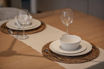 Elegant table setting for two. Minimalist white dinnerware, wine glasses, and wicker placemats on a wooden table. Dining concept.