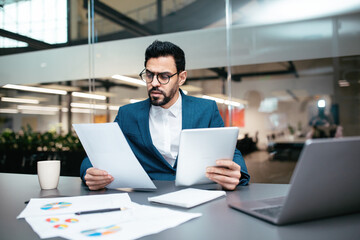 A business professional in a suit sits at a desk in a contemporary office, examining documents and analyzing charts while working on a laptop.
