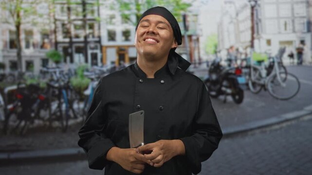 Young hispanic man chef in black uniform holding cleaver and smiling, hand gripping handle on cobblestone street; pride craftsmanship tradition.