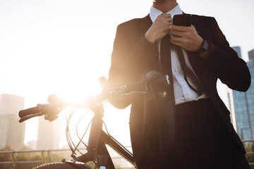 Modern businessman checking phone and adjusting tie by bicycle with city skyline, conveying professional ambition and eco-friendly commuting in a dynamic urban setting