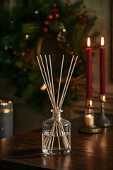 A Christmas interior scene showcases an elegant transparent glass reed diffuser with light wooden sticks on a dark wooden table. A softly glowing wreath of pine, red berries, and golden leaves is illu