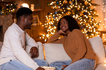 Smiling young African American husband and wife sit on a sofa in their living room, enjoying...
