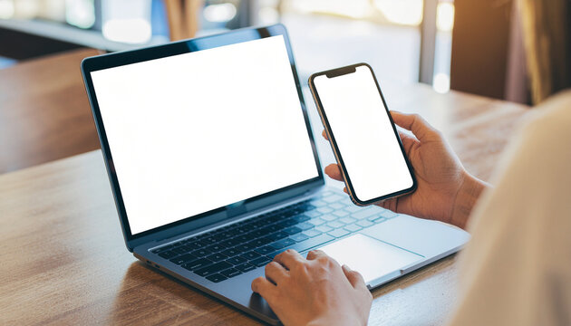 A close-up of a person using a laptop and holding a smartphone, both with blank white screens, on a wooden table. Perfect for showing work, mockups, or app use. - Powered by Adobe