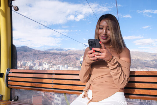Young woman with dark skin on the cable car in La Paz, Bolivia, using her cell phone with her hands - concept of technology