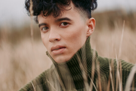 An authentic portrait of a young person in a knit turtleneck, captured in natural light amid tall grass, conveying realism and quiet intensity.