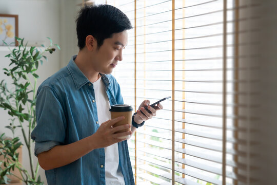 Happy Asian man drinking hot coffee in the morning using the phone while standing in the living room at home. Handsome young man walking around with a cup of hot drink, looks at a smartphone - Powered by Adobe