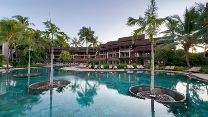 Beautiful resort swimming pool with teal water and palm trees growing inside, against a large hotel building backdrop at dusk