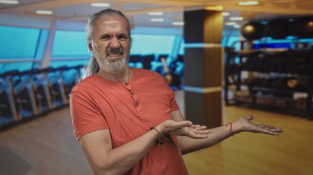 Man middle age with long grey hair grimacing, palms out presenting bare hands in gym sports center; disgust confusion.