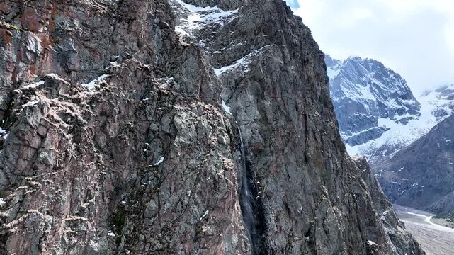 Close view of a thin meltwater stream flowing down a massive, dark gray, fractured granite rock face in an alpine environment