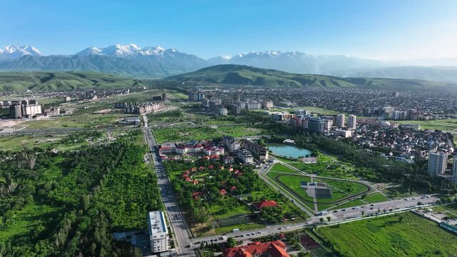 Wide aerial view of a modern area of the Bishkek city with apartment buildings, a central park, roads, and lush green foothills leading to the snow-capped Tian Shan mountains