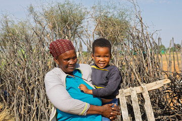 portrait old african woman holding child, village life, standing in front of fence from branches