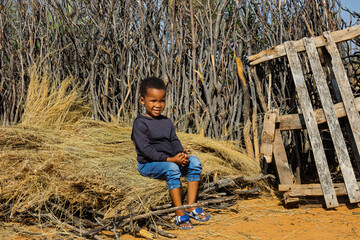 african village life, child sitting on thatched grass in the yard in front of the wood branches fence