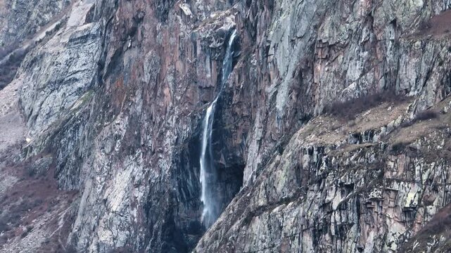 Close-up view of a tall, narrow waterfall plunging down a massive, sheer cliff face with highly textured, multicolored rock in a mountain gorge