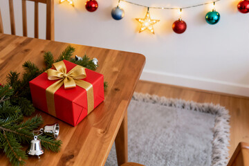 A bright red Christmas gift box with a golden ribbon is set on a wooden table, surrounded by pine branches and small silver bells, with holiday lights in the blurred background.